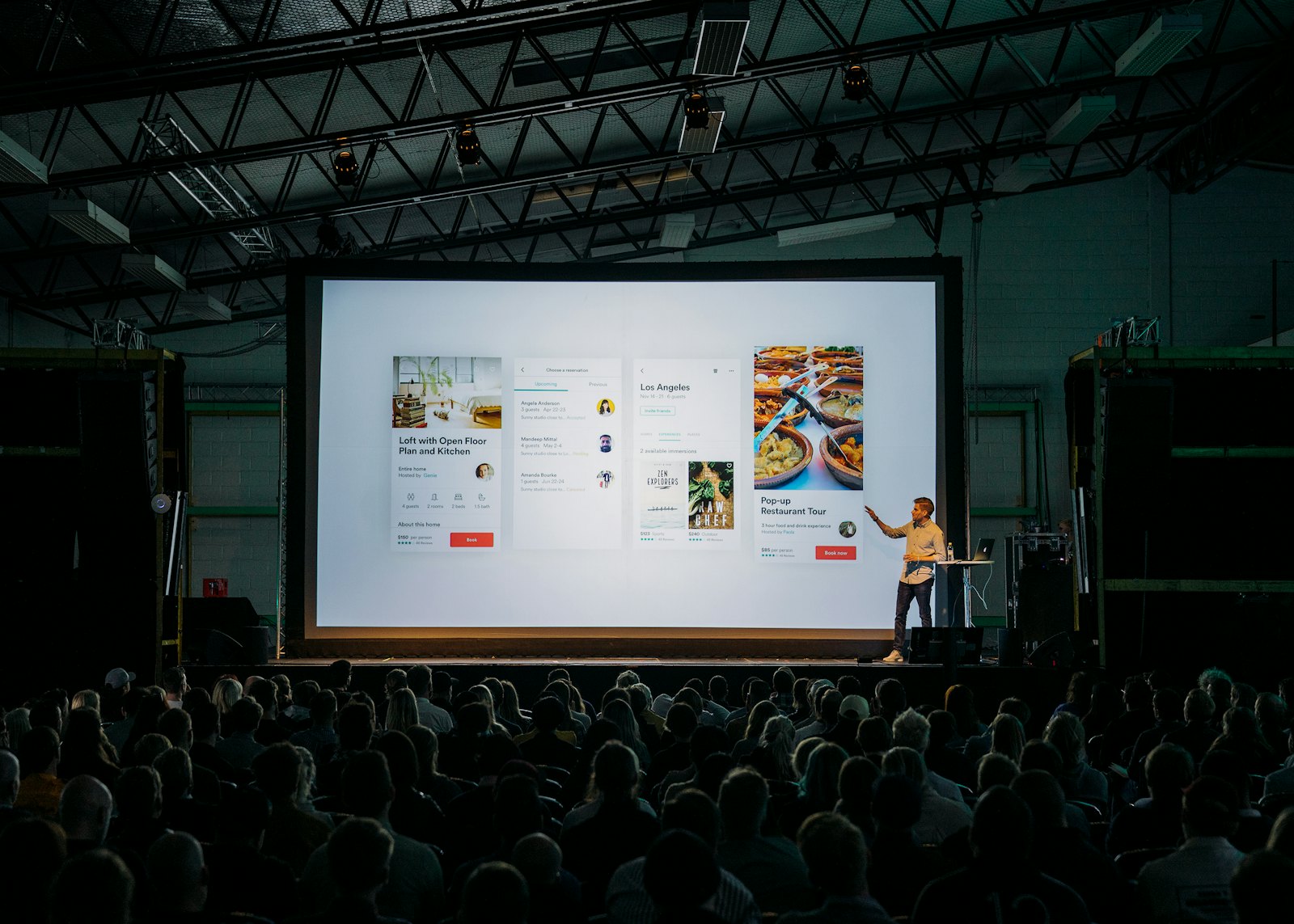 A presenter on stage in front of a large projection screen and an audience